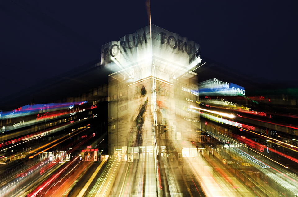 Byggnad med texten "Forum" och "Första" på fasaden, upplyst mot en mörk kvällshimmel, omgiven av ljusspår från rörelse och neonljus.