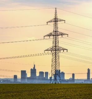Augstsprieguma elektrolīnijas tornis lauka vidū uz pilsētas panorāmas fona.