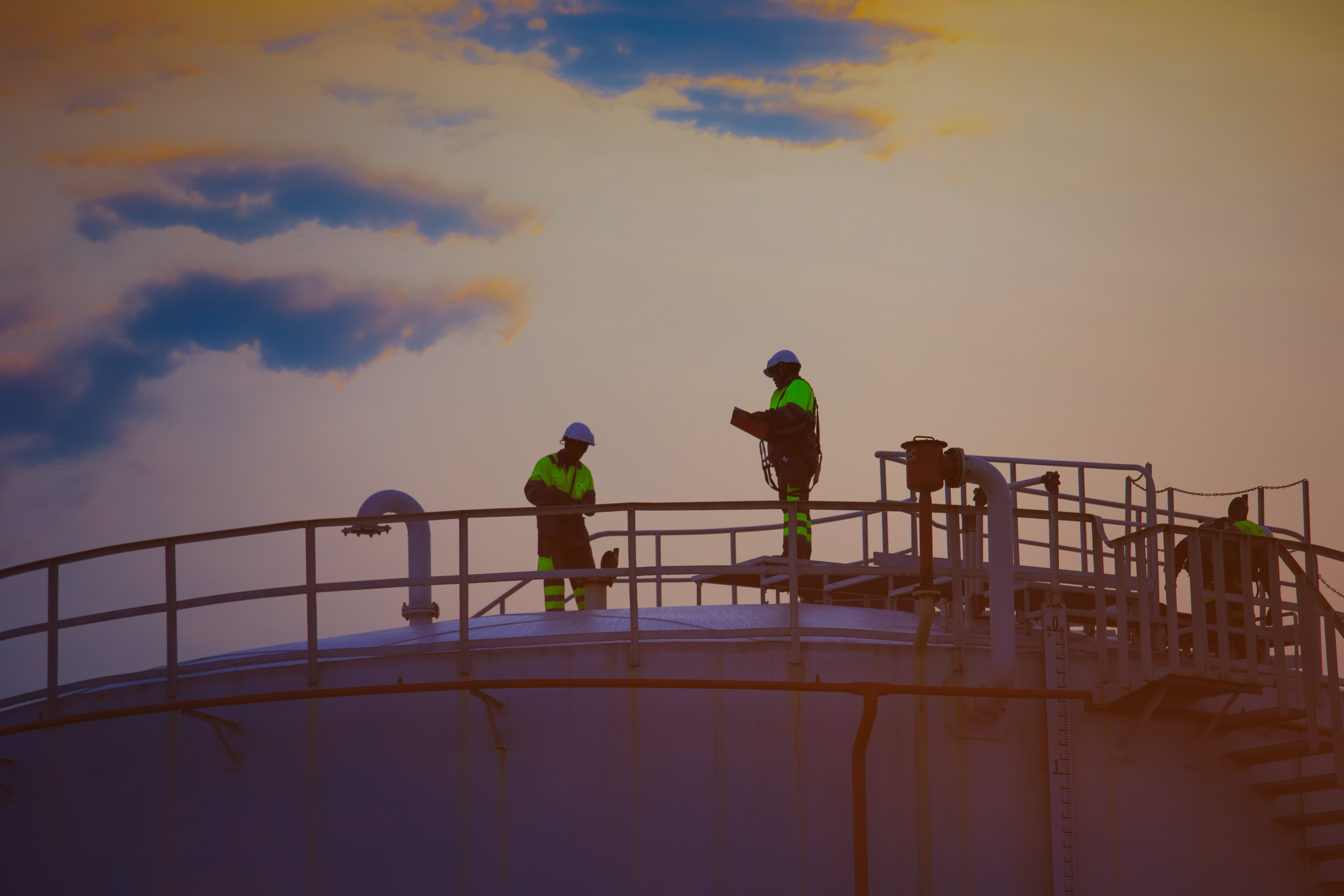 Industriālie darbinieki spilgtā drošības apģērbā uzglabāšanas tvertnes augšpusē saulrieta laikā.