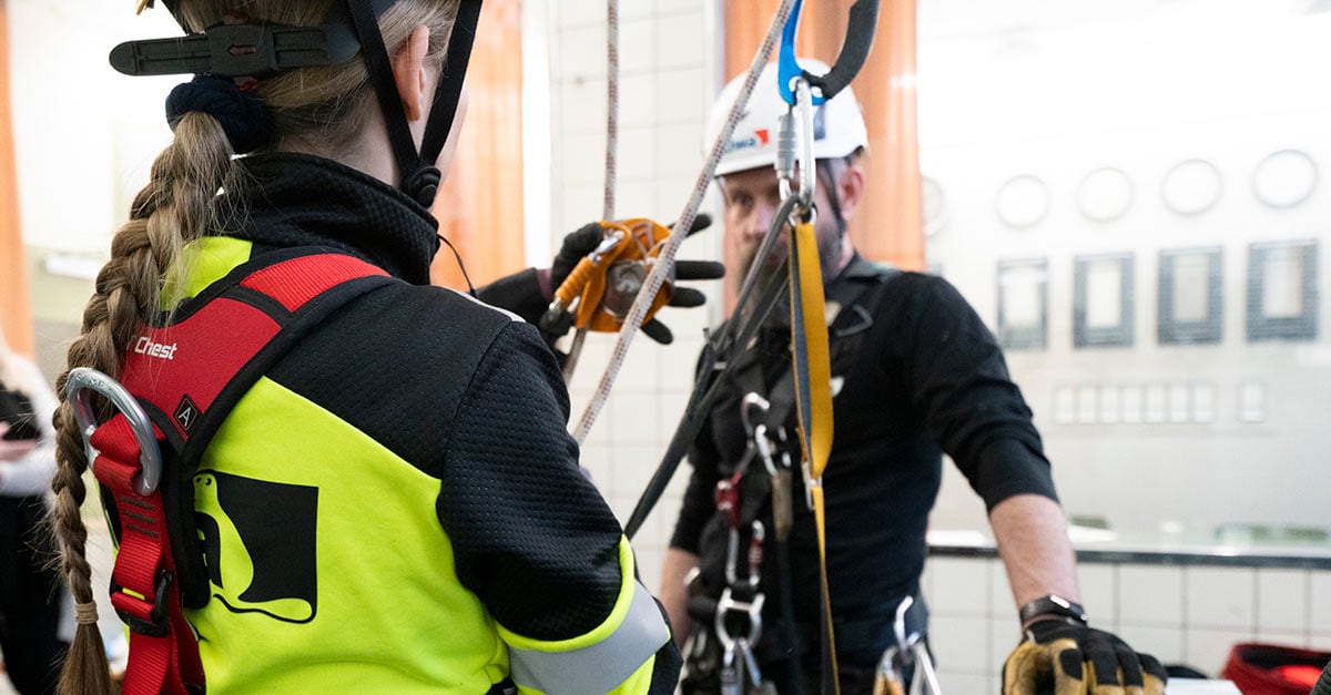 Cilvēki drošības ekipējumā piedalās industriālās alpīnisma nodarbībās telpā ar lieliem logiem.