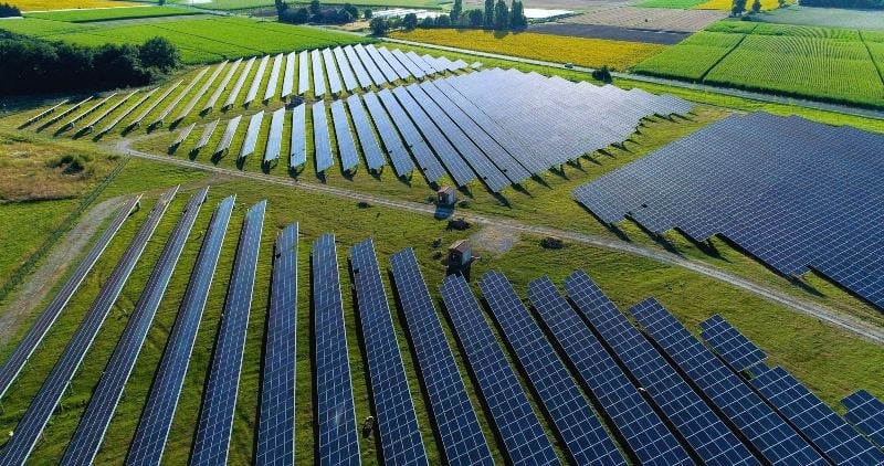 Aerial view of solar panels arranged in rows on green fields, surrounded by agricultural land.