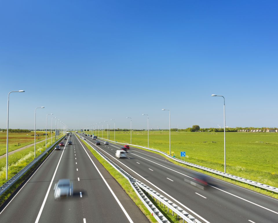 Autos rijden op een snelweg met meerdere rijstroken omgeven door groene weilanden onder een blauwe lucht.