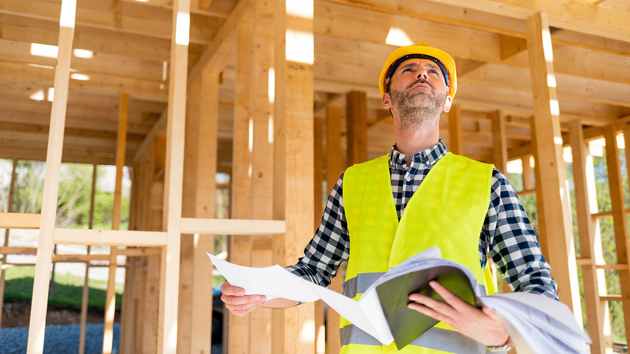 Manlig byggprojektledare i skyddshjälm och varselväst står med dokument i händerna framför en byggkonstruktion i trä.